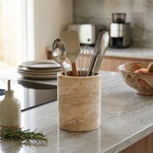 Beige marble utensil holder with stainless steel kitchen tools, including a whisk and spatula, on a marble countertop.