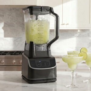 Professional black high-speed blender filled with a frozen green smoothie, standing next to margarita glasses with lime garnishes.