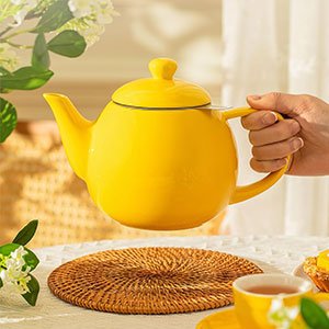 Bright yellow ceramic teapot being held over a woven rattan placemat in a sunlit kitchen setting.