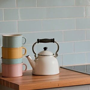 Classic white stovetop kettle next to a stack of colorful pastel mugs on a wooden countertop with light blue tile backsplash