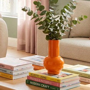 Vibrant orange bulbous ceramic vase with eucalyptus branches on a wooden coffee table.