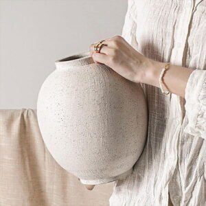 Close-up view of a person wearing an off-white linen shirt, holding a large, round textured white ceramic vase. The background is a clean grey wall.