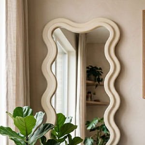 Large wavy-framed full-length floor mirror in a creamy white finish, leaning against a neutral wall near a green plant.